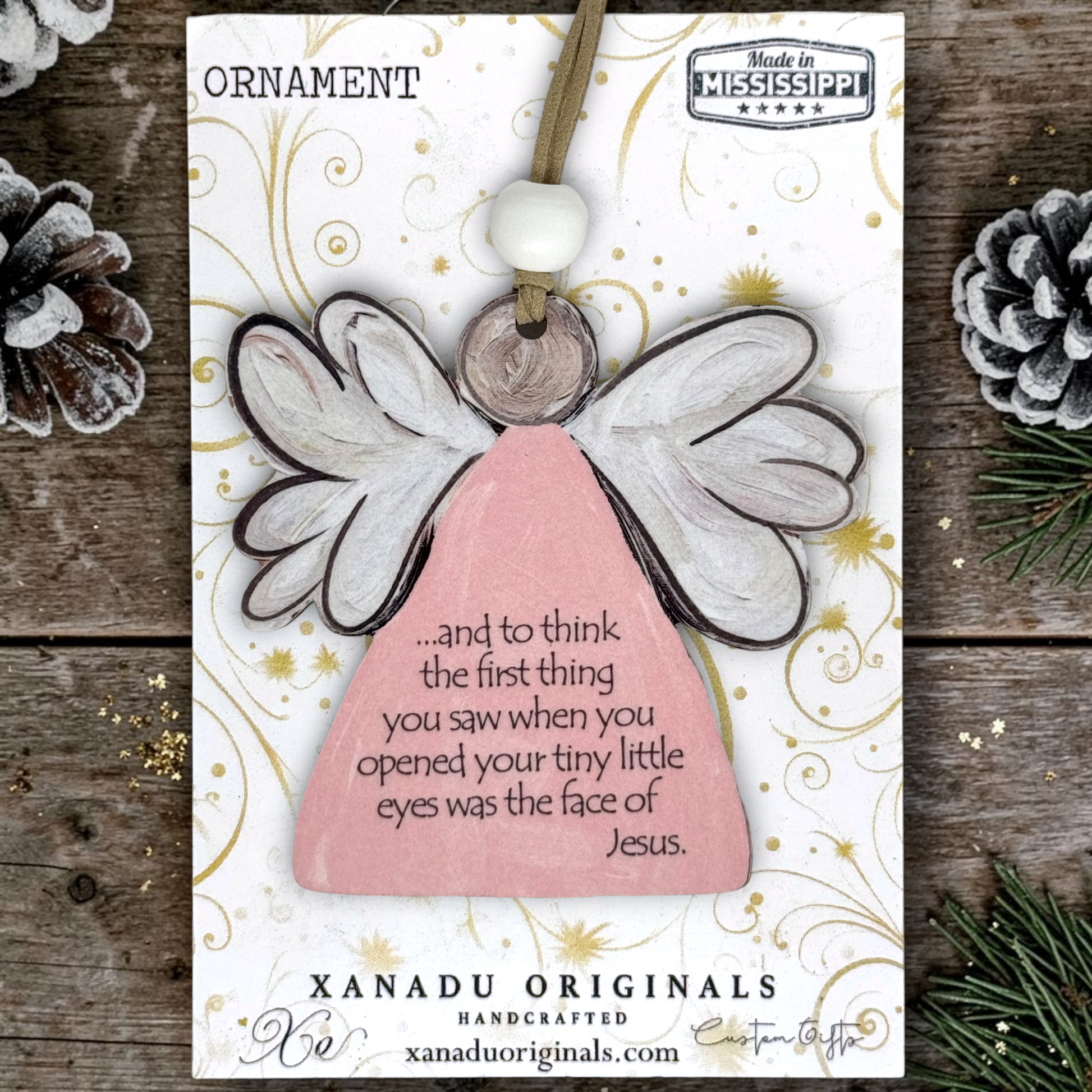A wooden angel ornament on a printed display card, photographed flat on a rustic wood surface with pinecones and evergreen branches around it. The ornament has a pale pink body, white wings with dark outlines, a white bead, and a small wooden disc.