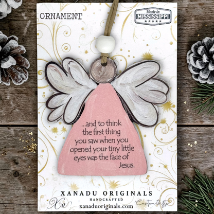 A wooden angel ornament on a printed display card, photographed flat on a rustic wood surface with pinecones and evergreen branches around it. The ornament has a pale pink body, white wings with dark outlines, a white bead, and a small wooden disc.