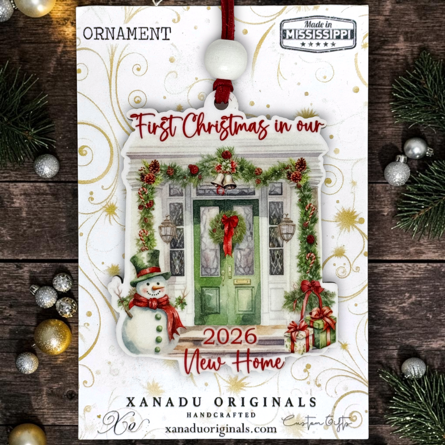 A Christmas ornament package from Xanadu Originals featuring a festive front door illustration with greenery, red lettering, a snowman, and the text 'First Christmas in our 2026 New Home.' The ornament hangs from a red cord with a white bead, shown against a dark wood background with pine branches and silver/gold ornaments around it.