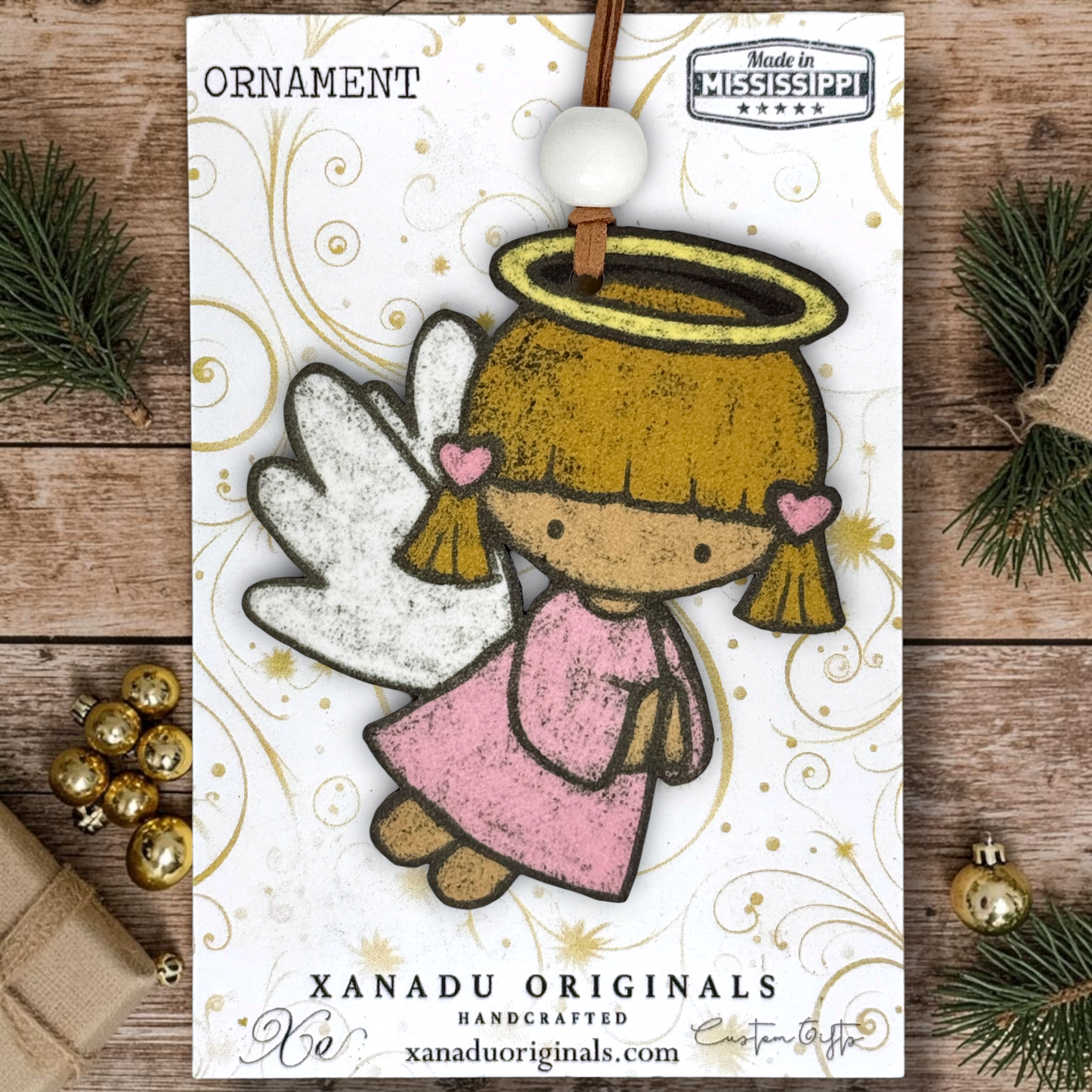 A handcrafted Christmas angel ornament by Xanadu Originals, featuring a whimsical angel with white wings, a gold halo, and a pink dress, shown on a wooden surface with holiday decorations.