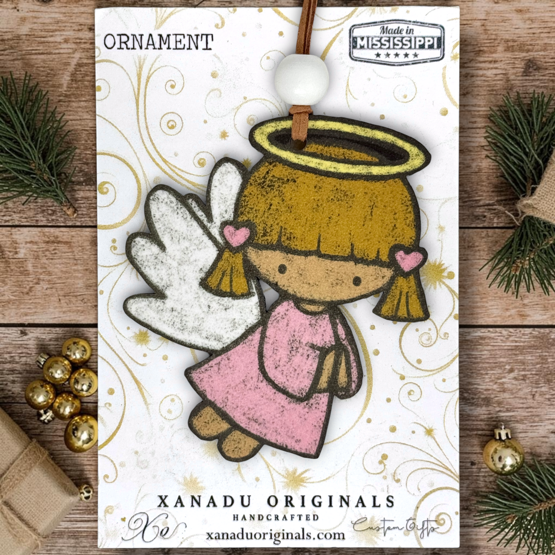 A handcrafted Christmas angel ornament by Xanadu Originals, featuring a whimsical angel with white wings, a gold halo, and a pink dress, shown on a wooden surface with holiday decorations.