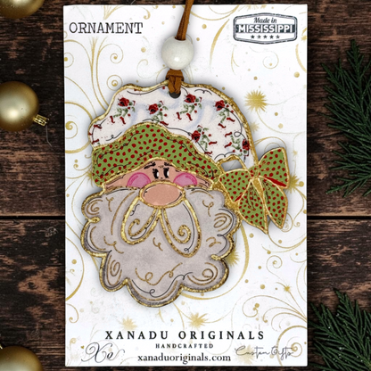 A handmade Christmas ornament card featuring a Santa Claus face ornament with a patterned hat, green polka-dot bow, and white beard, displayed on a branded card over a rustic wood background with holiday decorations.