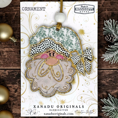 A handmade Christmas ornament card featuring a Santa Claus face ornament with a patterned hat, pink cheeks, and a white beard with gold glitter outlines. The card is branded 'Xanadu Originals' and includes a 'Made in Mississippi' seal, styled on a dark wood background with gold ornaments and pine branches.
