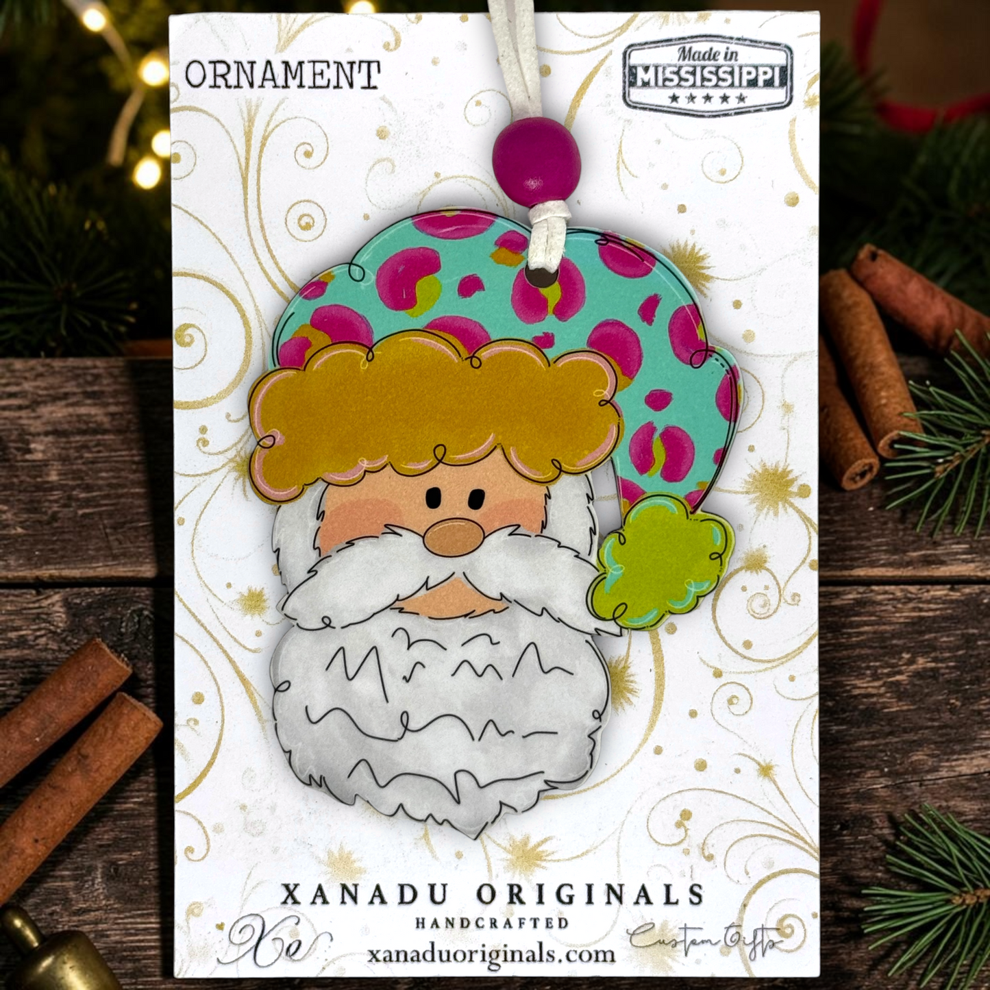 A handmade Christmas ornament featuring a stylized Santa Claus face with a white beard, gold hair, and a teal hat with pink and green spots, displayed on a branded card over a rustic holiday background.
