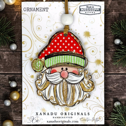 A festive Christmas ornament featuring a handcrafted Santa Claus face with a red polka-dot hat, green striped trim, white beard, and gold glitter details, hanging from a brown ribbon on a white card. The card is photographed on a dark wood surface with holiday greenery and ornaments around it.