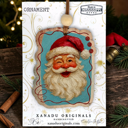 Handcrafted Christmas ornament featuring a smiling Santa Claus face on a turquoise background, mounted on a white branded display card. The card reads 'ORNAMENT' and 'XANADU ORIGINALS HANDCRAFTED xanaduoriginals.