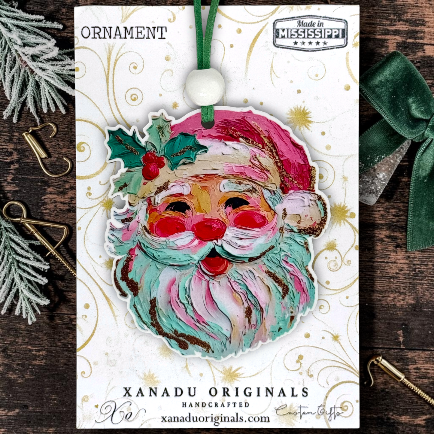 A festive handcrafted Christmas ornament featuring a smiling Santa face with a pink hat, mint green beard, and holly accent, displayed on branded Xanadu Originals packaging over a rustic dark wood background with pine branches and a green ribbon.
