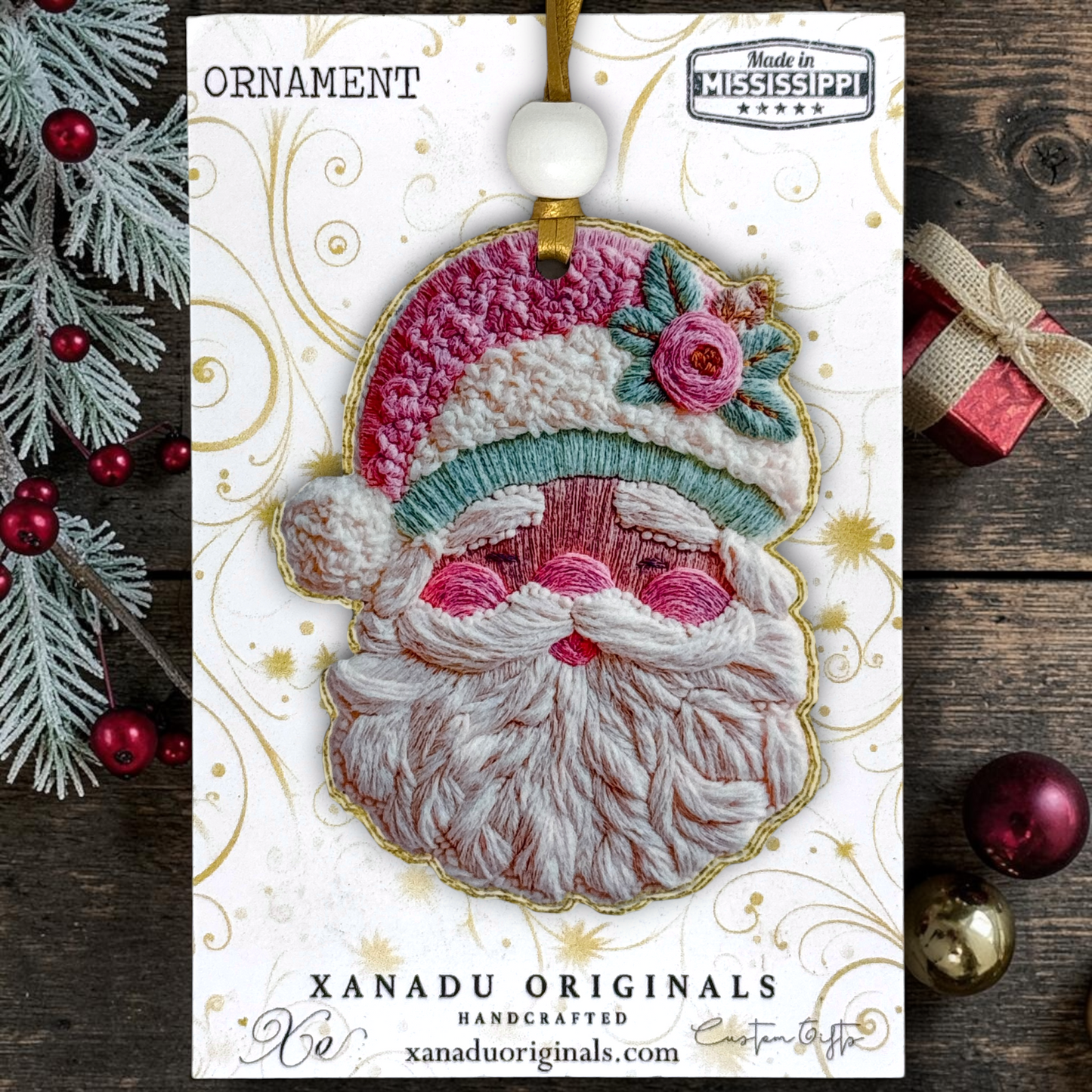A handmade embroidered Christmas ornament featuring a stylized Santa face with a pink and white hat, teal trim, and pink cheeks, displayed on a branded card over a rustic wood background with holiday decorations.
