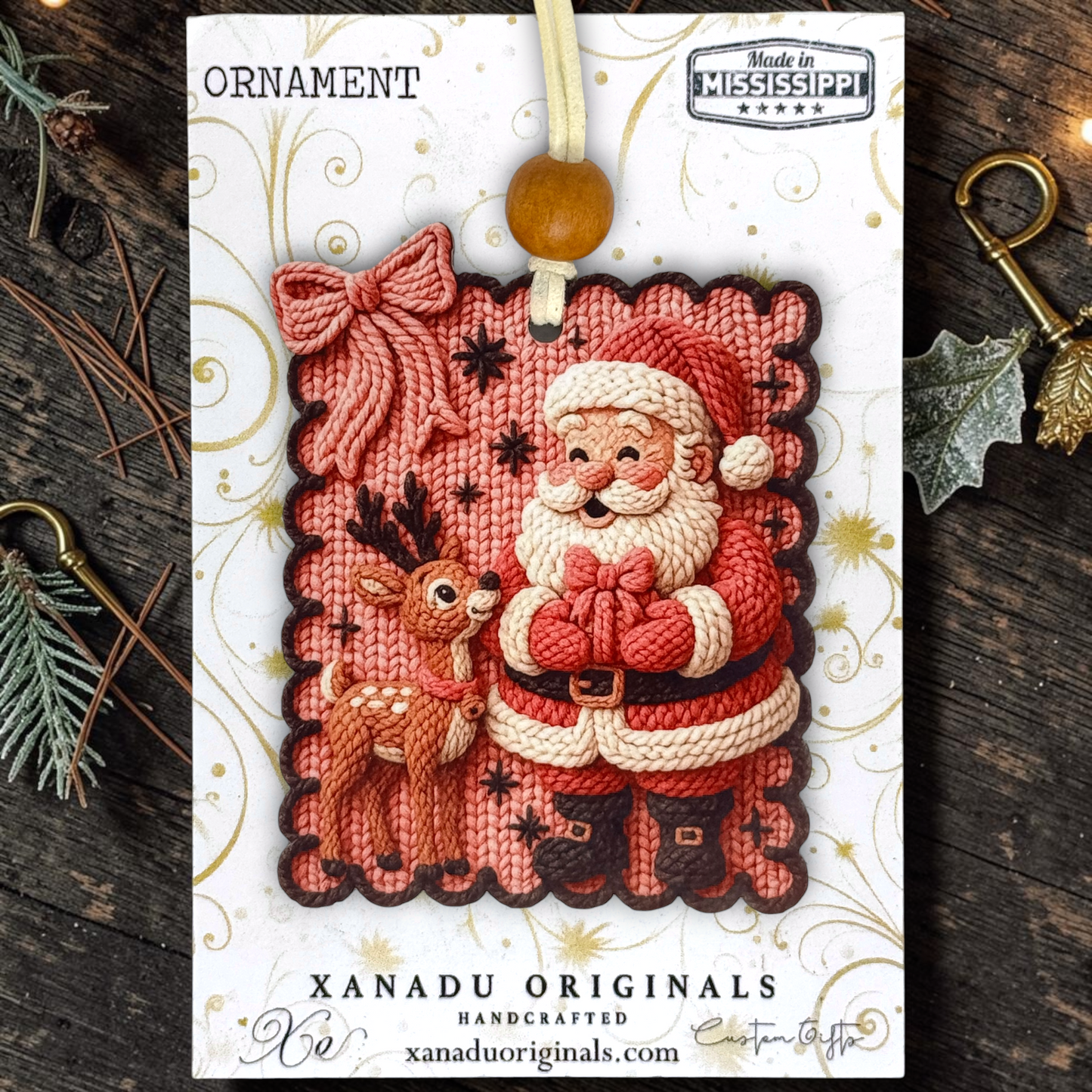 A Christmas ornament on a  card featuring a faux knitted Santa Claus and deer ornament, with a wooden bead hanging loop. The card reads 'ORNAMENT' and 'XANADU ORIGINALS HANDCRAFTED', photographed on a rustic dark wood surface with pine branches and decorative keys around it.