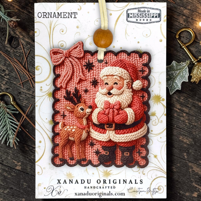 A Christmas ornament on a  card featuring a faux knitted Santa Claus and deer ornament, with a wooden bead hanging loop. The card reads 'ORNAMENT' and 'XANADU ORIGINALS HANDCRAFTED', photographed on a rustic dark wood surface with pine branches and decorative keys around it.