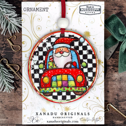 A round Christmas ornament card featuring a festive illustration of Santa driving a red car on a black-and-white checkered background. The ornament is displayed on a branded card from Xanadu Originals, surrounded by holiday props including pine branches, gold metal clips, and a green ribbon on a rustic wood surface.