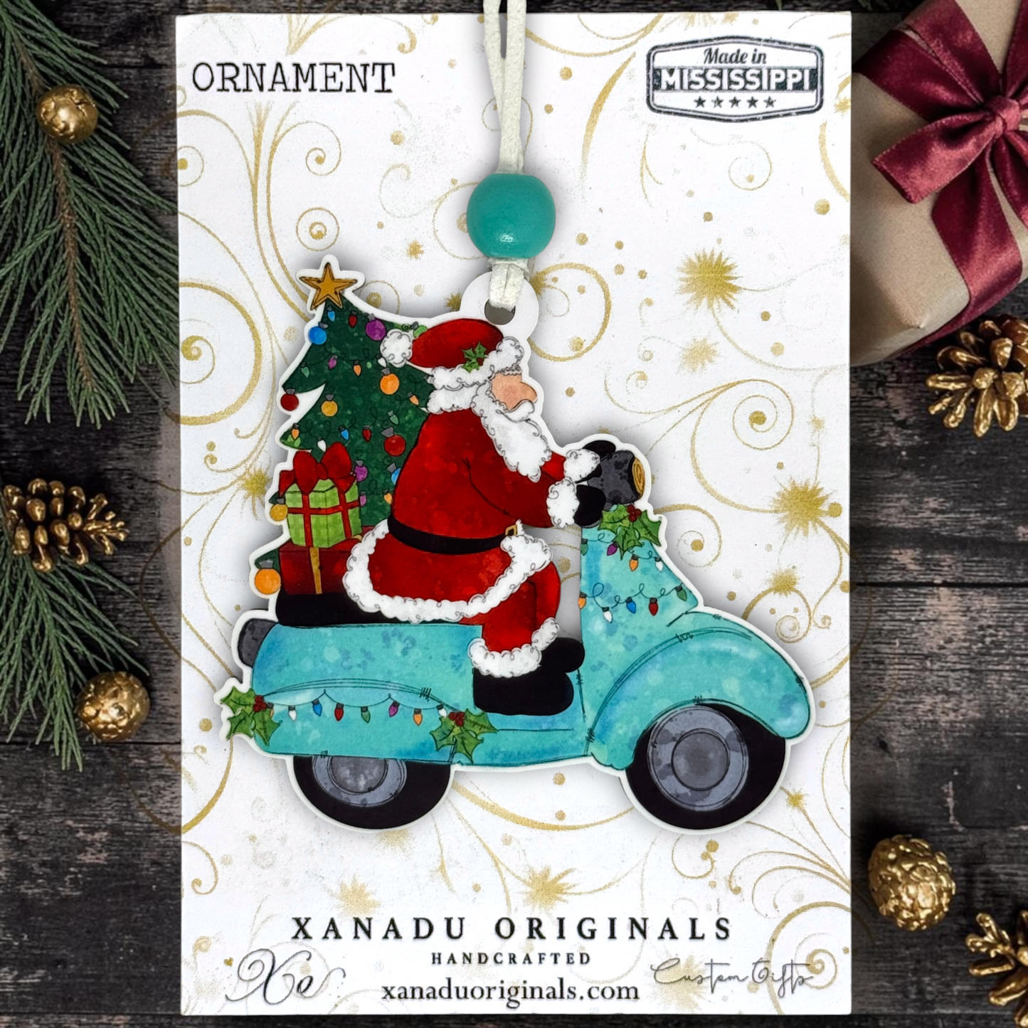 Handcrafted Christmas ornament featuring Santa Claus riding a turquoise scooter with a decorated tree and gifts, mounted on a decorative white card with gold swirls. The card is styled on a dark wood surface with pine branches, gold pinecones, and a wrapped gift.