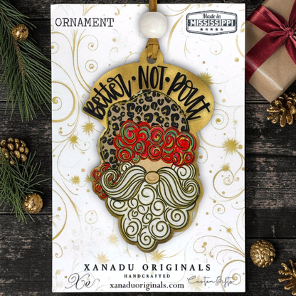 A leopard print "You Better Not Pout" Santa wood ornament displayed flat on a white Xanadu Originals packaging card featuring elegant gold swirl patterns.