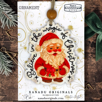 Handcrafted Christmas ornament by Xanadu Originals featuring an illustrated Santa Claus with the text 'Believe in the magic of Christmas' on a white card, styled on a rustic wooden surface with holiday props.