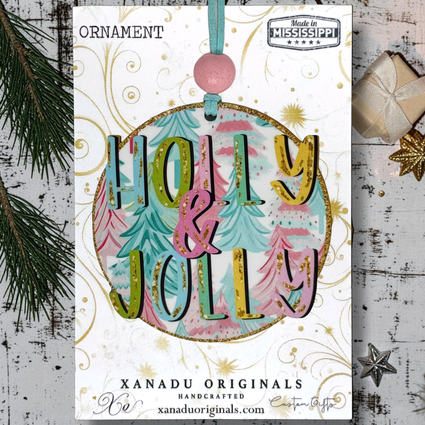 A Christmas ornament  on a card from Xanadu Originals featuring the words “Holly & Jolly” layered over pastel Christmas trees. The ornament has a gold glitter border, a pink hanging loop, and decorative gold swirls on a white card.