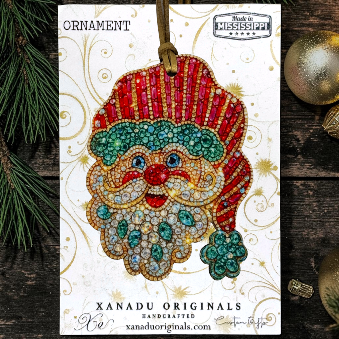 A festive Christmas ornament card featuring a Santa Claus face made of rhinestones, with the text 'ORNAMENT' and 'XANADU ORIGINALS HANDCRAFTED'. The card is styled on a dark wood surface with pine branches and gold ornaments.