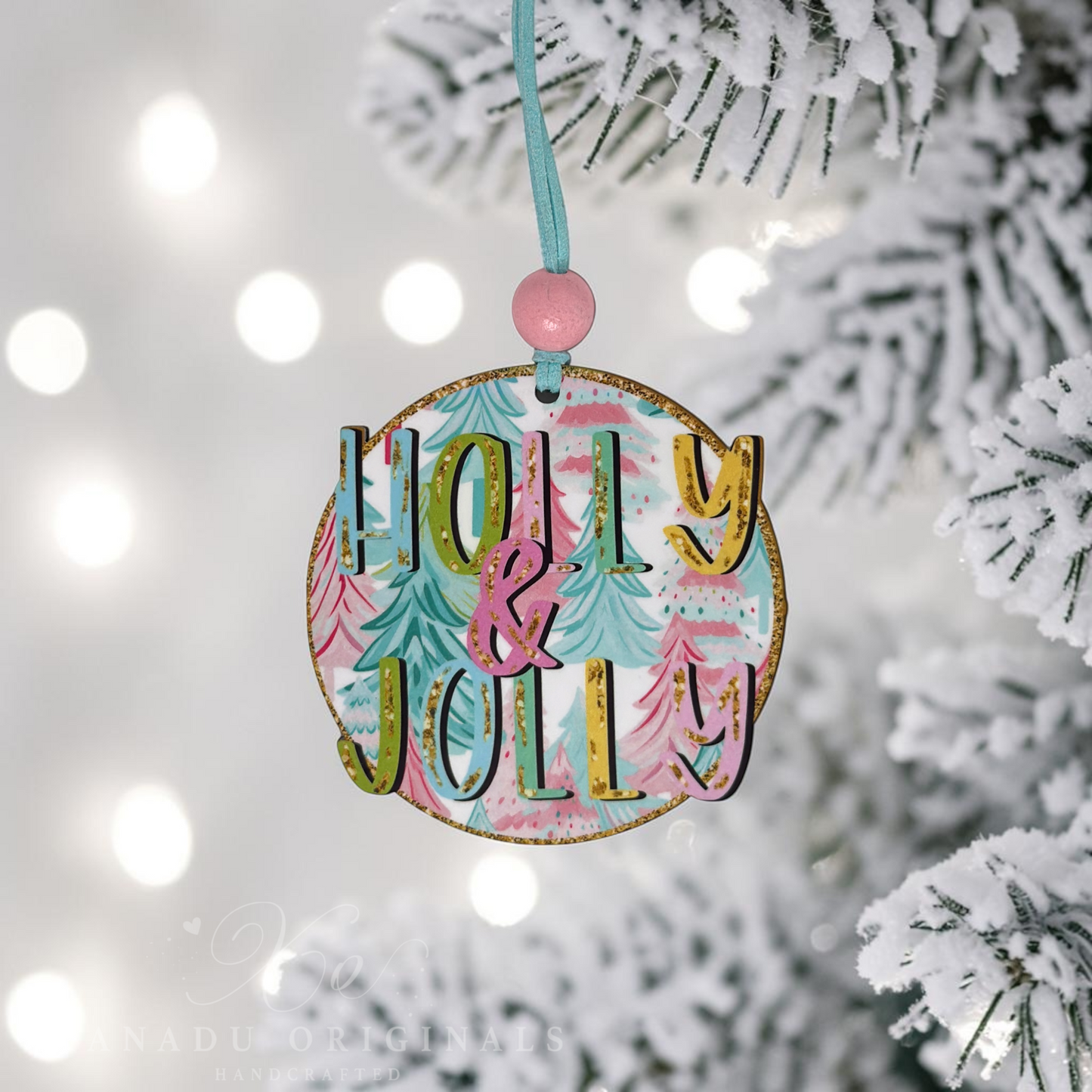 A round wooden Christmas ornament hanging from a light blue ribbon with a pink bead, featuring the words “Molly & Jolly” in gold glitter letters over a pastel tree pattern. The ornament is shown hanging from a snowy pine branch with soft white bokeh lights in the background.