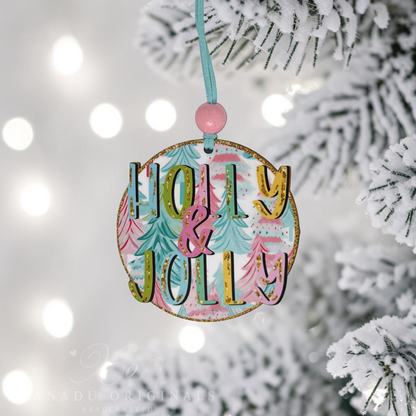 A round wooden Christmas ornament hanging from a light blue ribbon with a pink bead, featuring the words “Molly & Jolly” in gold glitter letters over a pastel tree pattern. The ornament is shown hanging from a snowy pine branch with soft white bokeh lights in the background.