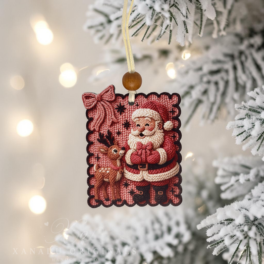 A rectangular Christmas ornament featuring a stitched Santa Claus and a reindeer, hanging from a yellow cord with a brown bead against a blurred snowy Christmas tree background with warm string lights.
