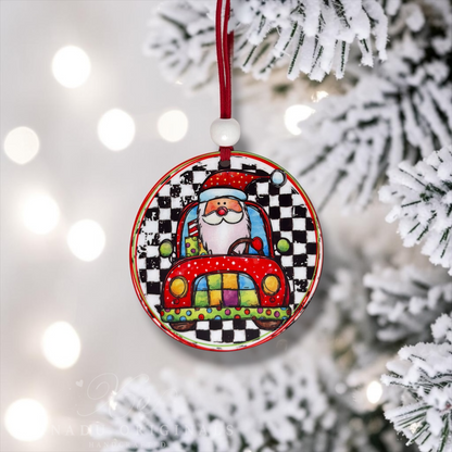 A round Christmas ornament featuring a colorful cartoon Santa driving a red car on a black-and-white checkered background, hanging from a red ribbon. The ornament is photographed in front of a blurred snowy Christmas tree with warm bokeh lights.