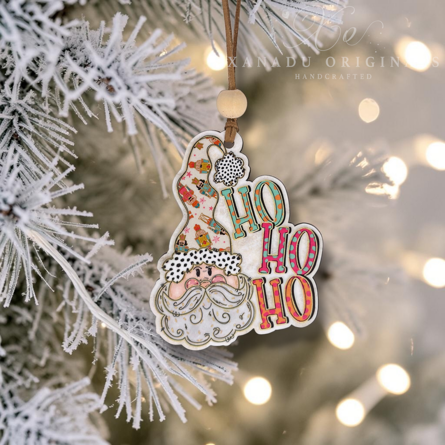 A festive Christmas ornament shaped like a Santa figure with the words “HO HO HO” in colorful patterned letters, hanging from a flocked pine tree branch with warm bokeh lights in the background. A watermark reading “XANADU ORIGIN HANDCRAFTED” appears near the top.