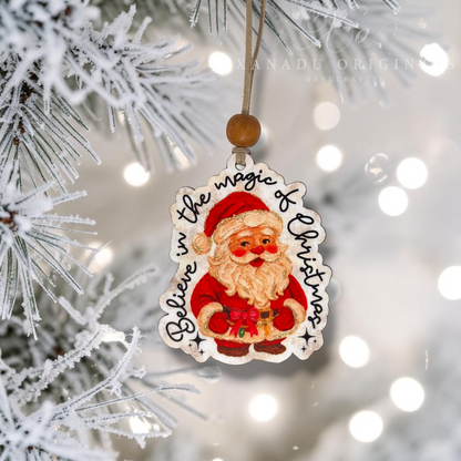 A Christmas ornament shaped like Santa Claus hanging from a frosted tree branch. The ornament features an embroidered Santa in a red suit with a white beard, surrounded by the words 'Believe in the magic of Christmas' in black script.