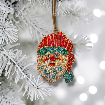 A handmade Christmas ornament shaped like Santa's face and hat, decorated with red, green, gold, and white rhinestones, hanging from a brown string on a white frosted Christmas tree branch with soft bokeh lights in the background. A watermark for 'XANADU ORIGIN HANDCRAFTED' is visible near the top.