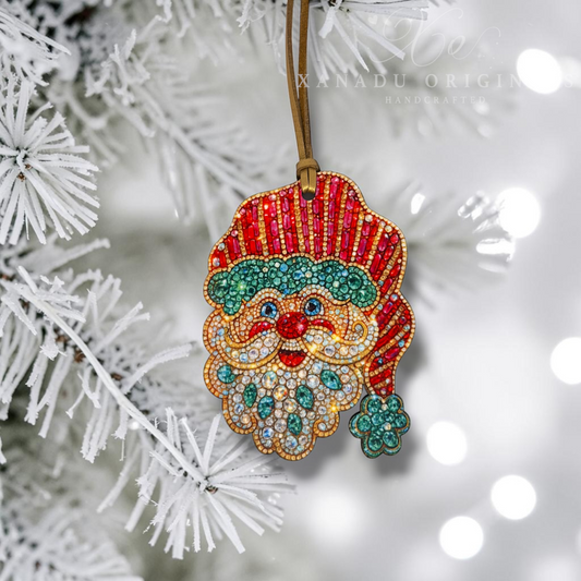A handmade Christmas ornament shaped like Santa's face and hat, decorated with red, green, gold, and white rhinestones, hanging from a brown string on a white frosted Christmas tree branch with soft bokeh lights in the background. A watermark for 'XANADU ORIGIN HANDCRAFTED' is visible near the top.