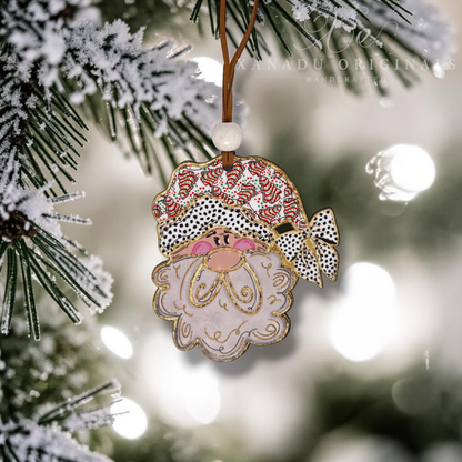 A Santa Claus Christmas ornament hanging from a snowy pine tree branch, with a warm bokeh background of lights.