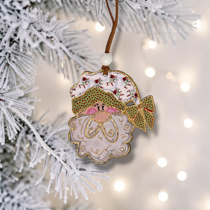 A decorative Christmas ornament shaped like a gnome or Santa face hangs from a white-frosted tree branch. The ornament has a white face and beard, green patterned hat and bow, pink cheeks, and gold embroidered details.