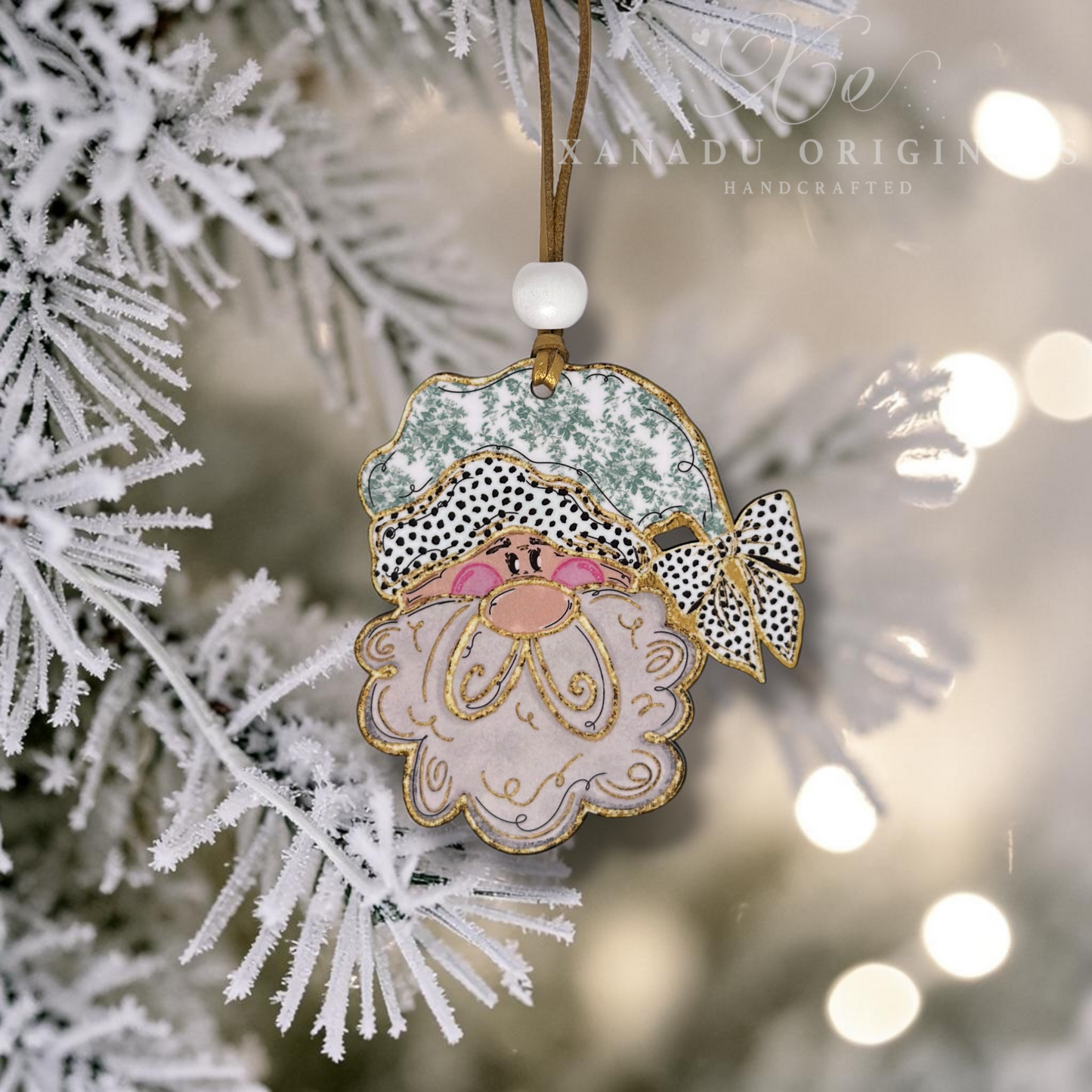 A handcrafted Santa Claus Christmas ornament hanging from a white frosted Christmas tree branch. The ornament features a light pink beard, a black and white spotted hat with a green patterned top, gold glitter outlines, and a brown hanging string with a white bead.