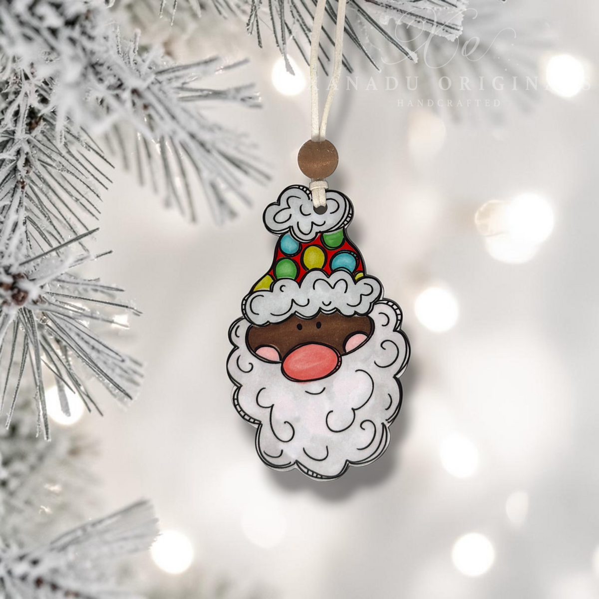 A dark skin tone whimsical Santa wood ornament with a red and green polka dot hat and dark wood bead hanging from a snowy white flocked Christmas tree branch with warm bokeh background lights.