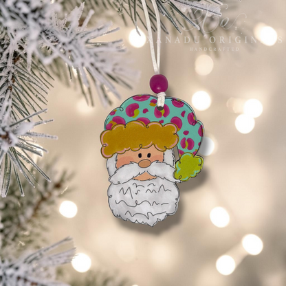 A festive handcrafted Christmas ornament shaped like a Santa figure with a white beard, yellow hair, and a teal hat with pink and green spots, hanging from a snowy pine branch against a warm bokeh background of holiday lights. A watermark reading 'Xanadu Originals Handcrafted' is visible in the upper right.