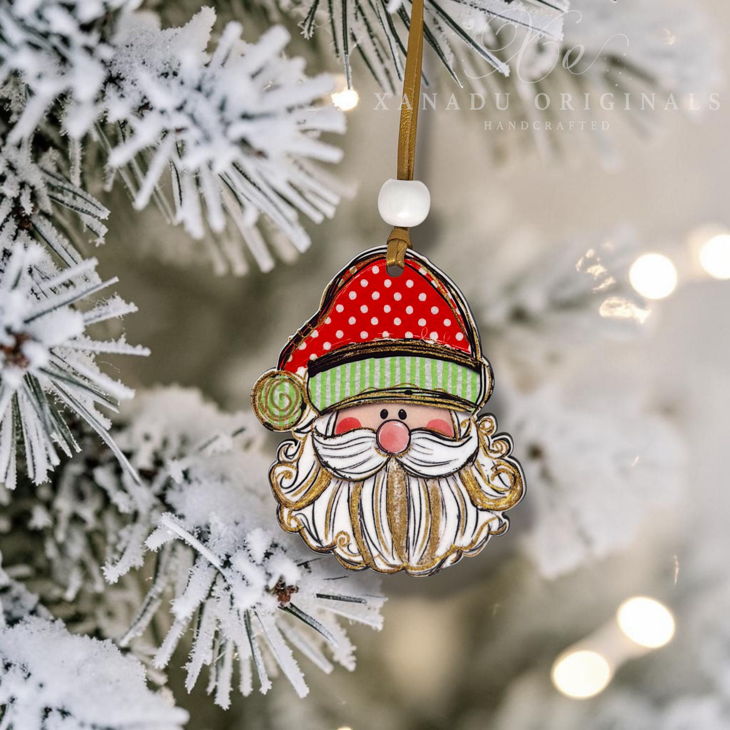 A handcrafted Christmas ornament shaped like a Santa face hanging from a snowy pine tree branch. The ornament features a red hat with white polka dots, a green striped band, a pink nose, and a white beard with gold detailing.