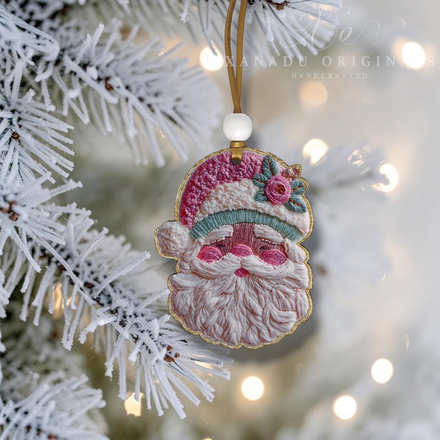 An embroidered Christmas ornament shaped like Santa's face hangs from a white frosted Christmas tree branch. The ornament features pink and white embroidery for Santa's beard and cheeks, a red and white hat with a green leaf accent, and a gold embroidered outline.