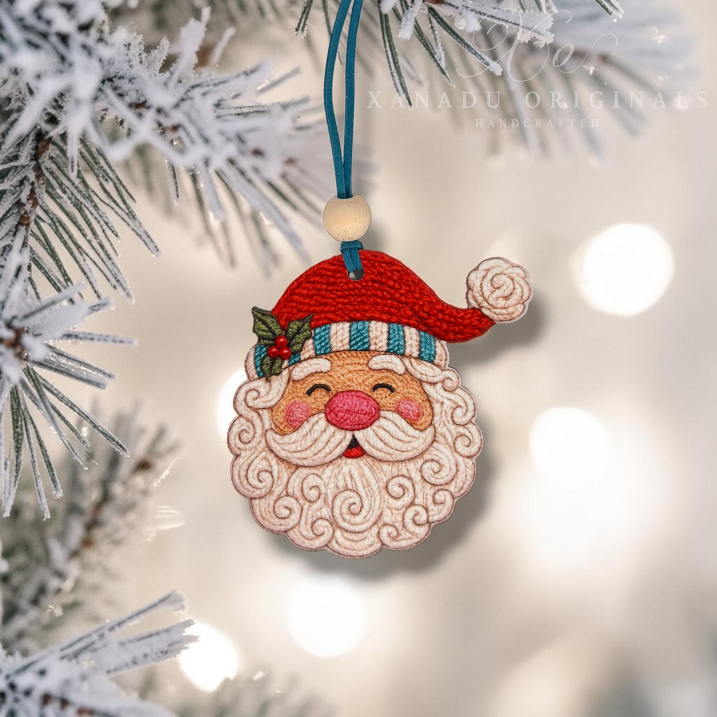 A handmade embroidered Christmas ornament shaped like Santa's face, hanging from a blue ribbon on a frosted pine branch. The ornament features a red hat, a striped blue and white hatband, a pink nose, and a curly white beard.