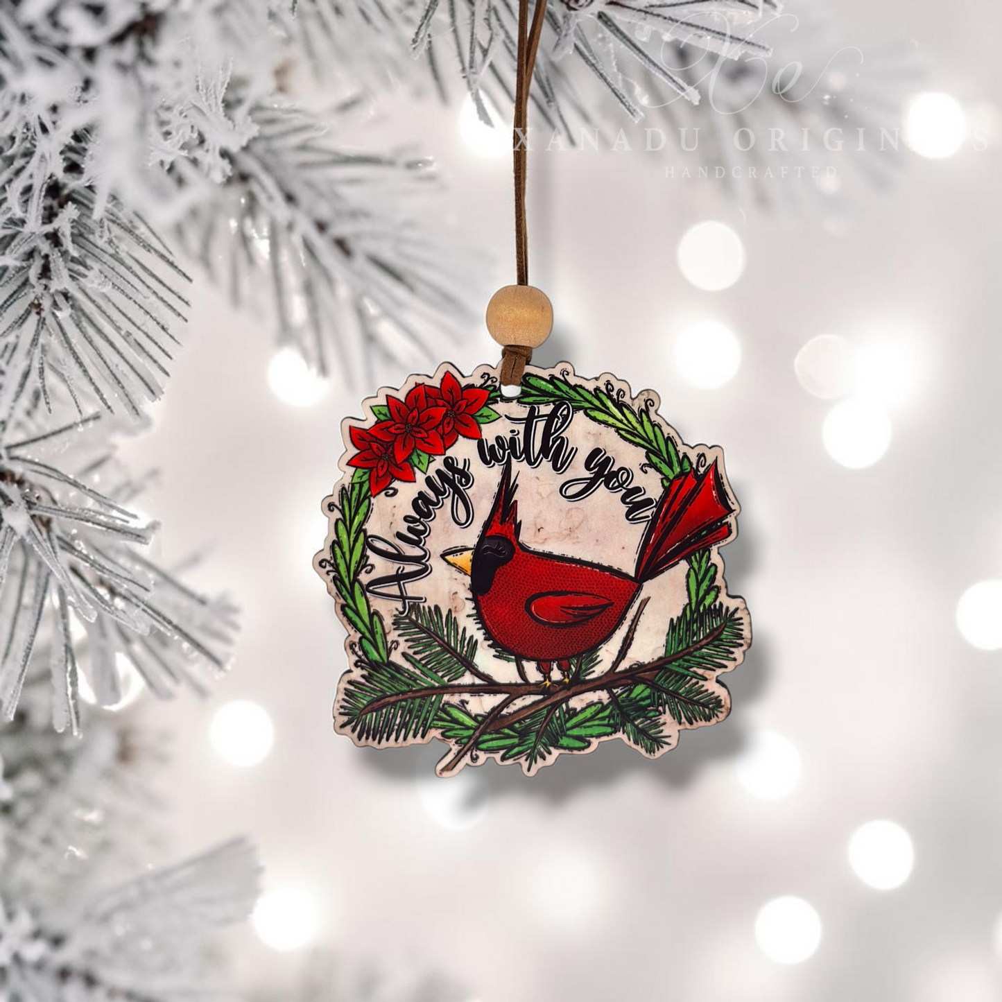 A handcrafted wooden Christmas ornament featuring a red cardinal perched on a wreath with red poinsettias and the words 'Always with you,' hanging from a tree branch against a soft white bokeh background.