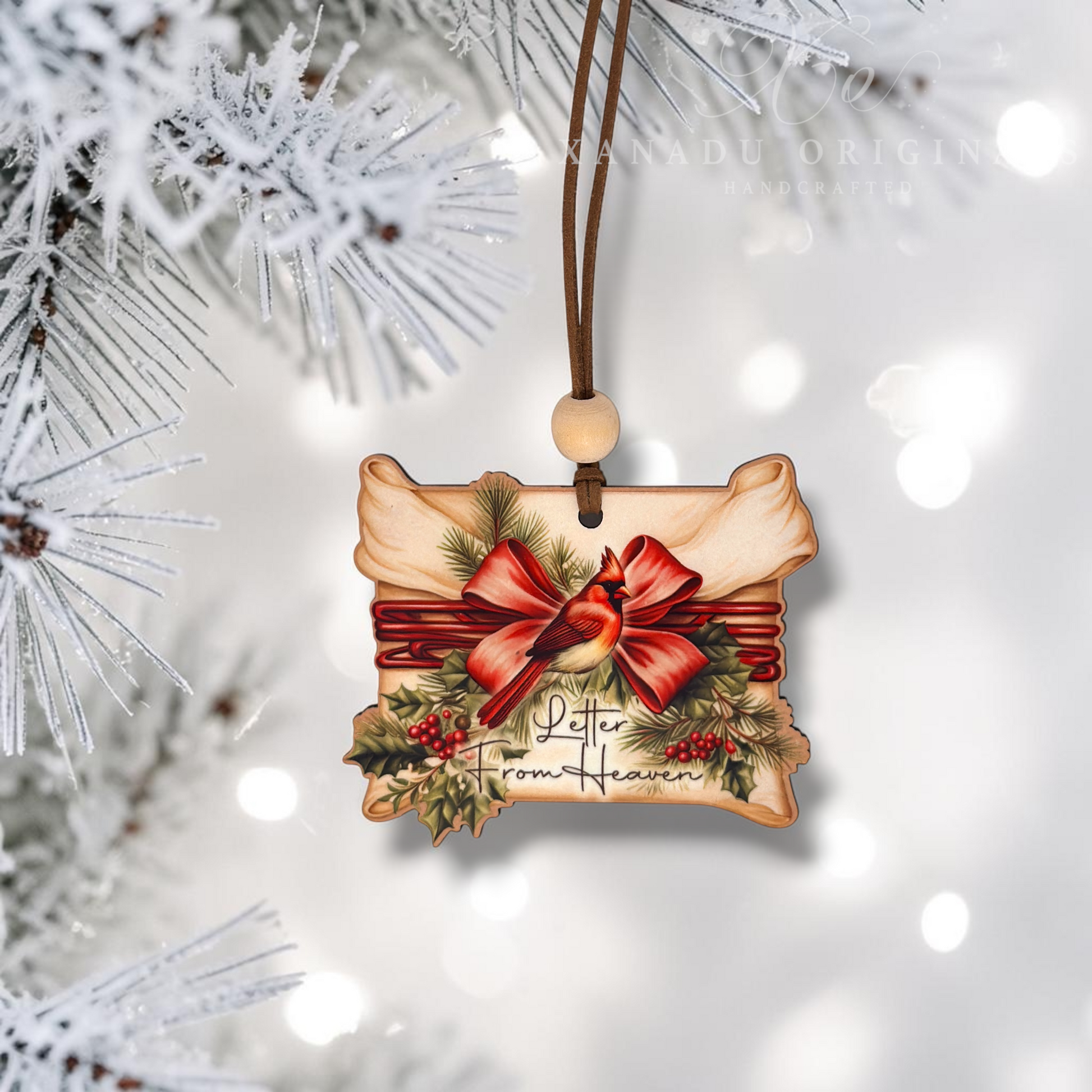 A rectangular wooden Christmas ornament with a printed holiday design featuring a red bird, red bow, green holly leaves, and the words 'Peter from Heaven,' hanging from a brown cord with a wooden bead. The ornament is shown against a soft white bokeh background with blurred snowy tree branches around the edges.