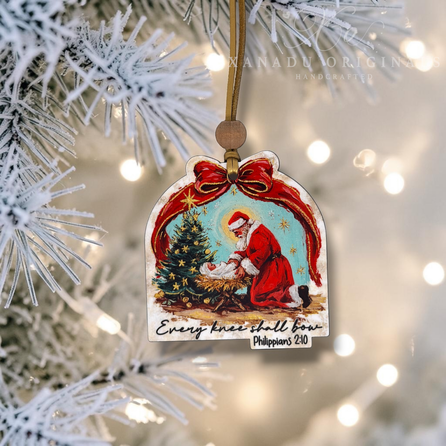 Handcrafted wooden Christmas ornament hanging on a frosted pine tree branch with warm string lights in the background. The ornament features a painted nativity scene with Santa, a baby Jesus, and a Christmas tree, with the text 'Every knee shall bow' and 'Philippians 3:20'.