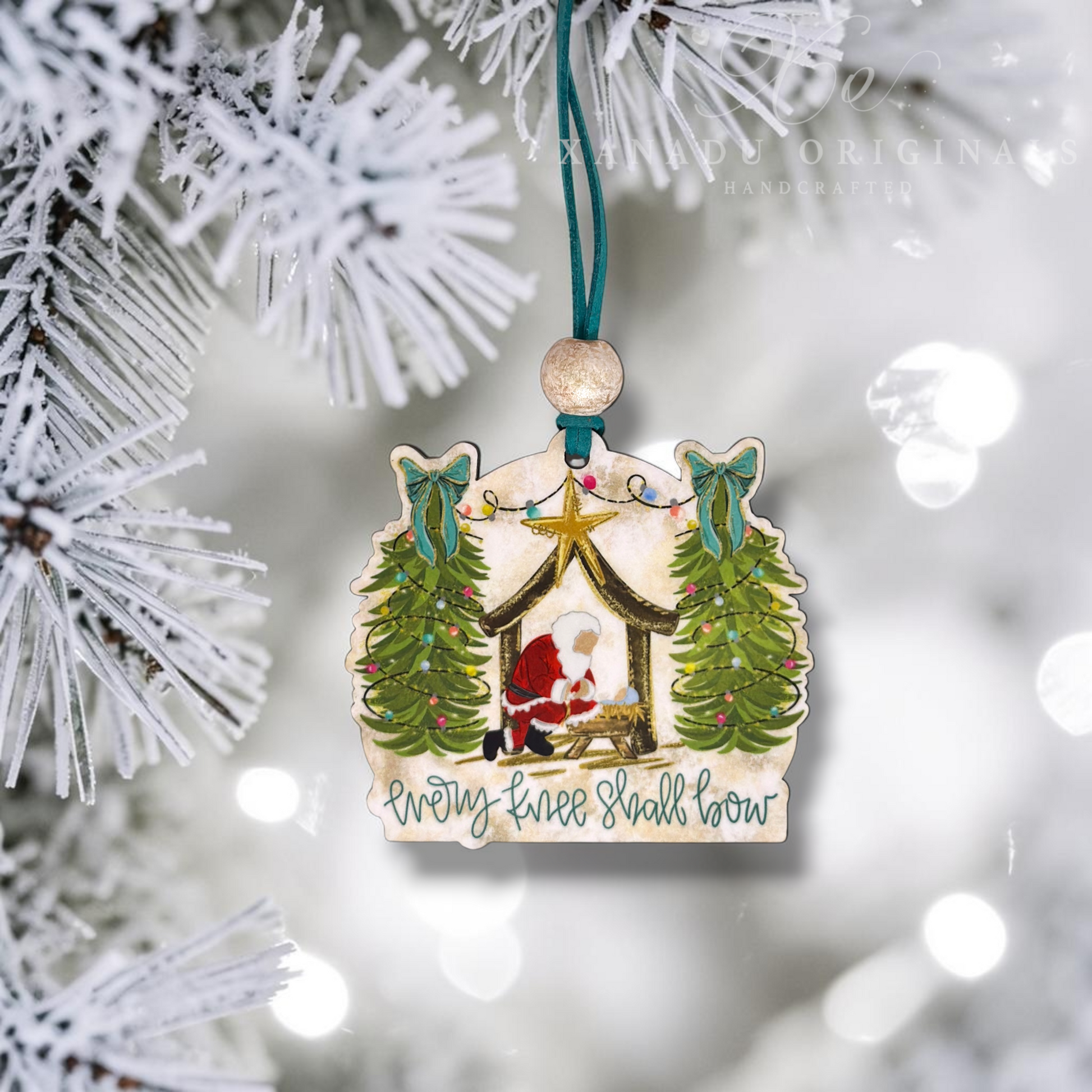 A handcrafted wooden Christmas ornament shaped like a stable, hanging from a teal ribbon on a frosted Christmas tree branch. The ornament features an illustrated scene of the baby Jesus in a manger, flanked by two decorated Christmas trees, with the text 'Every knee shall bow' written below.