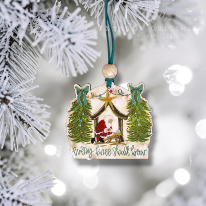 A handcrafted wooden Christmas ornament shaped like a stable, hanging from a teal ribbon on a frosted Christmas tree branch. The ornament features an illustrated scene of the baby Jesus in a manger, flanked by two decorated Christmas trees, with the text 'Every knee shall bow' written below.