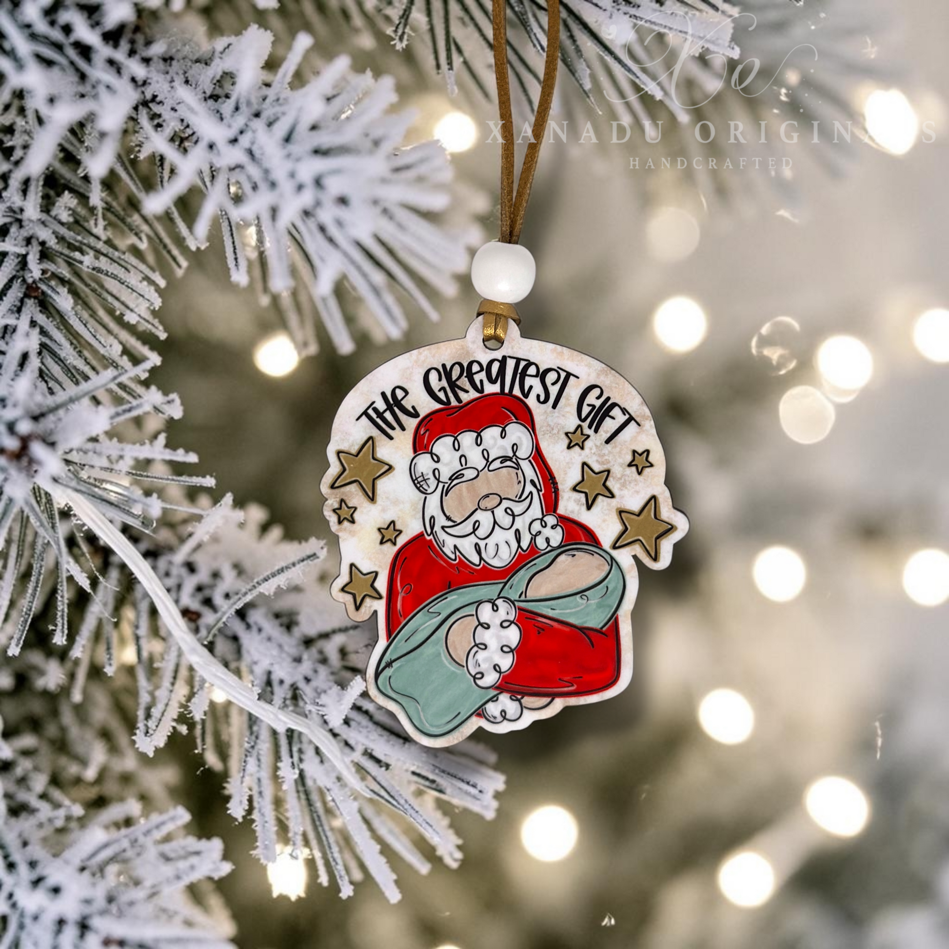 A handcrafted wood Christmas ornament featuring an illustration of Santa Claus holding a sack with the text 'THE GREATEST GIFT' above him, hanging from a brown ribbon on a frosted Christmas tree branch with warm bokeh lights in the background.