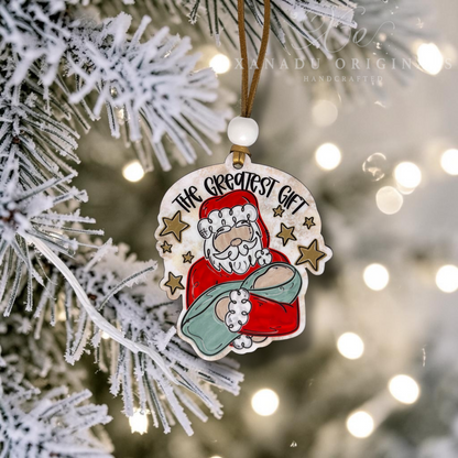 A handcrafted wood Christmas ornament featuring an illustration of Santa Claus holding a sack with the text 'THE GREATEST GIFT' above him, hanging from a brown ribbon on a frosted Christmas tree branch with warm bokeh lights in the background.