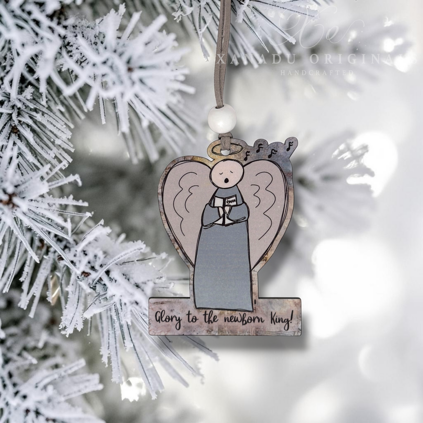A heart-shaped wooden Christmas ornament featuring a hand-drawn angel with a halo and the text 'Glory to the newborn King!' hanging from a flocked pine tree branch.
