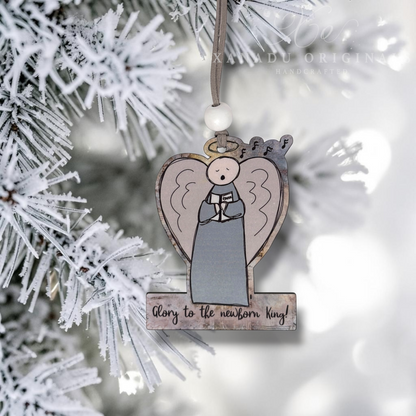 A heart-shaped wooden Christmas ornament featuring a hand-drawn angel with a halo and the text 'Glory to the newborn King!' hanging from a flocked pine tree branch.