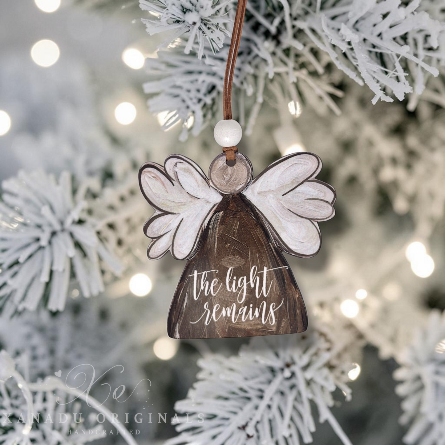 A wooden angel Christmas ornament hanging from a brown string, featuring white wings, a white bead, and a brown angel with the words 'the light remains' in white script. The ornament is photographed against a blurred flocked Christmas tree with warm bokeh lights.
