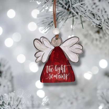 A handcrafted angel Christmas ornament hanging from a frosted pine branch. The ornament features a red angel with the words 'the light remains' in white script, white wings with dark outlines, a brown hanging cord, and a white bead.