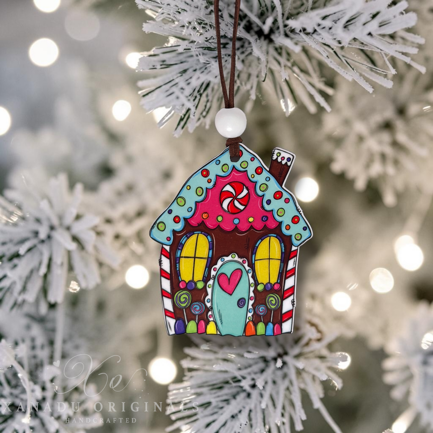 A colorful gingerbread house Christmas ornament hanging from a white bead on a brown string, set against a blurred white frosted Christmas tree background with warm bokeh lights.