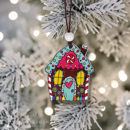A colorful gingerbread house Christmas ornament hanging from a white bead on a brown string, set against a blurred white frosted Christmas tree background with warm bokeh lights.