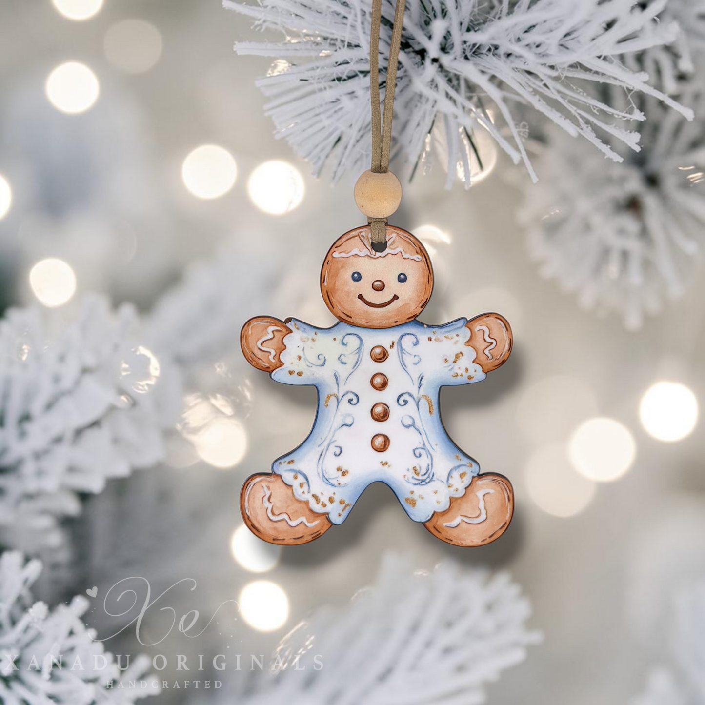 A gingerbread man Christmas ornament hanging from a white frosted tree branch, with soft bokeh lights in the background. The ornament is tan with a light blue icing dress and small gold accents.